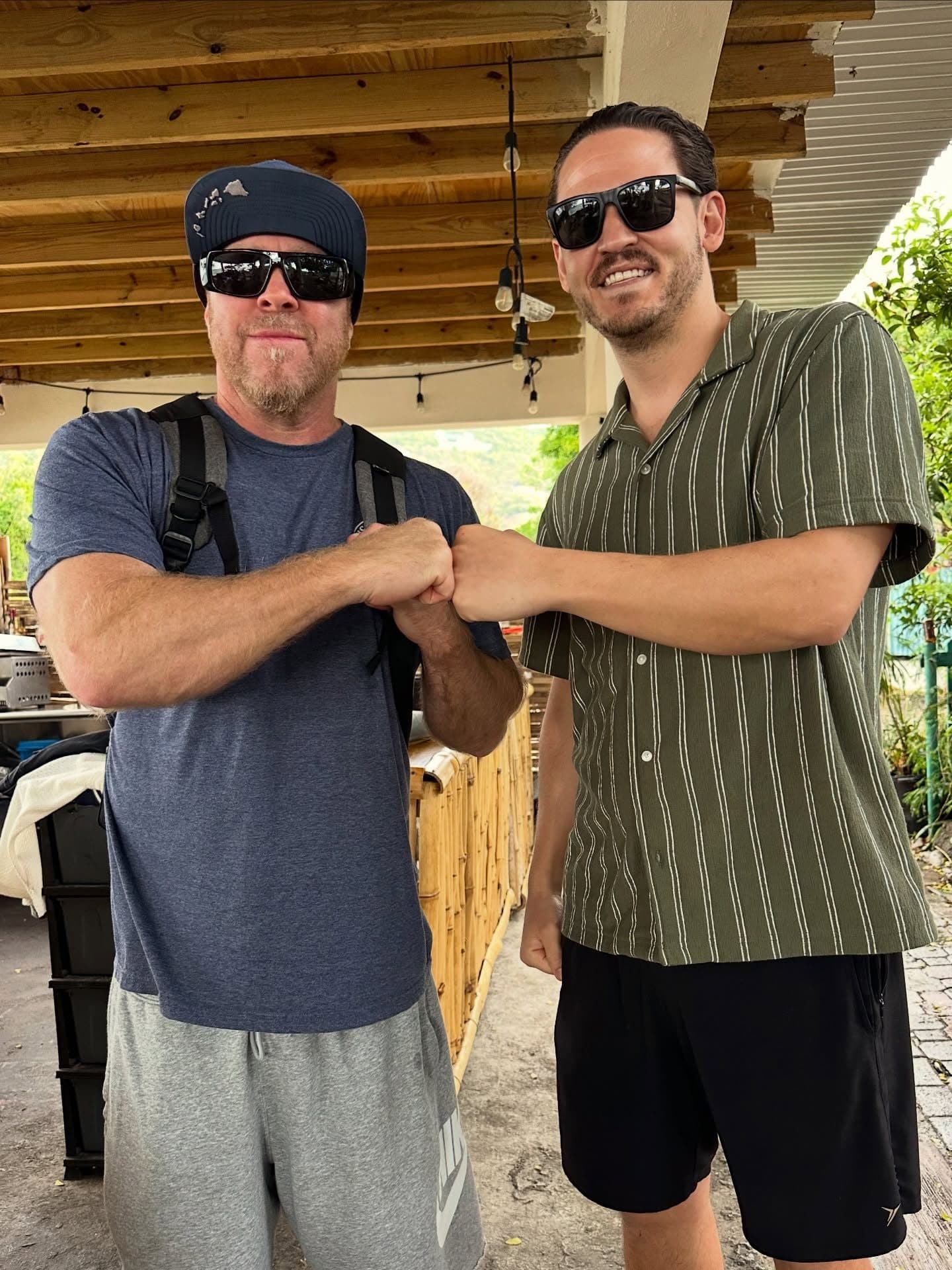 DJ Maxafari and a friend bumping fists under a wooden bar roof